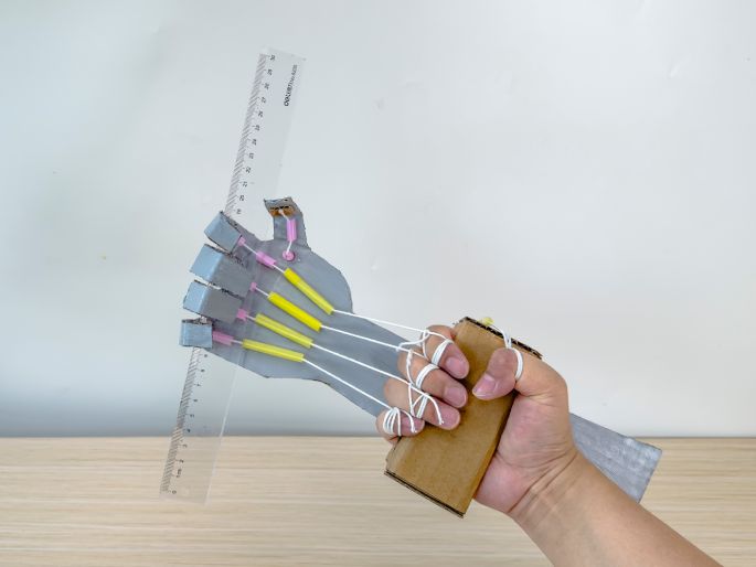 A hand operates a DIY cardboard robotic hand, gripping a clear plastic ruler against a light-colored background. The robotic hand is constructed from silver-painted cardboard, with pink and yellow straws along the fingers. White strings connect the fingers to a small cardboard box worn on the back of the hand, serving as the control mechanism. The hand is demonstrating the robotic hand's ability to grasp and hold an object. This image likely illustrates a science or engineering project demonstrating principles of robotics.