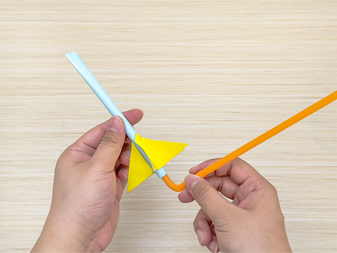 Two hands are assembling a toy rocket or projectile. The left hand holds a pale blue paper straw with a bright yellow triangular fin attached. The right hand holds a flexible orange straw and is inserting it into the open end of the blue straw. The background is a light woodgrain surface.