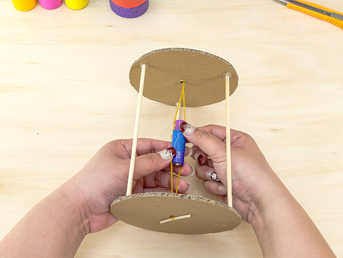Two hands hold a small ballista or catapult constructed from cardboard, wooden dowels, and rubber bands. A small battery, wrapped in blue textured material, is held in place between the two cardboard discs by a yellow rubber band, which appears to be the launching mechanism. The hands, with decorated fingernails, are positioning the battery. The construction sits on a light wooden surface, and colorful craft supplies are visible in the background, indicating a DIY or crafting project.