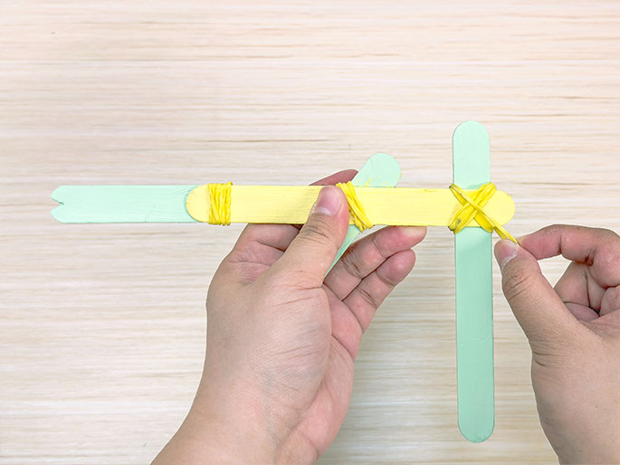 A hand holds and fires a small, toy gun made from popsicle sticks. The gun is primarily light green, with a yellow barrel and trigger mechanism. Yellow rubber bands secure the sticks together, and a thin blue stick serves as the projectile. The hand pulls back on the blue stick, launching it forward against a plain white background.