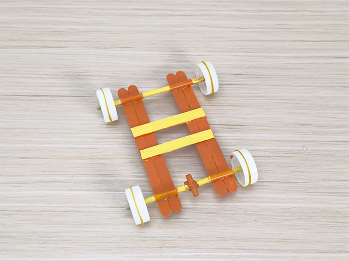 A small, simple car constructed from orange and yellow popsicle sticks sits on a light wooden surface. The car has four wheels, made from small white cylinders, likely cardboard or plastic. A rubber band is looped around the center of the car's frame, likely serving as the propulsion mechanism. The car is stationary, and the video provides a top-down view of its construction.