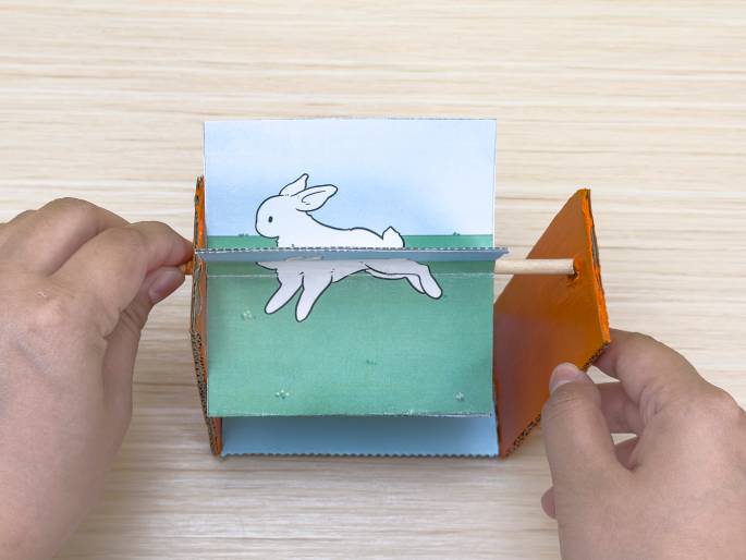Two hands hold a small zoetrope or animation device made of cardboard and paper. The central image depicts a white rabbit running against a light blue sky and green grass background. A wooden dowel is inserted through the sides of the device, presumably to spin the images and create the illusion of movement. The sides of the zoetrope are orange cardboard. The background is a light wood-grained surface. This image showcases a simple, handmade animation toy.