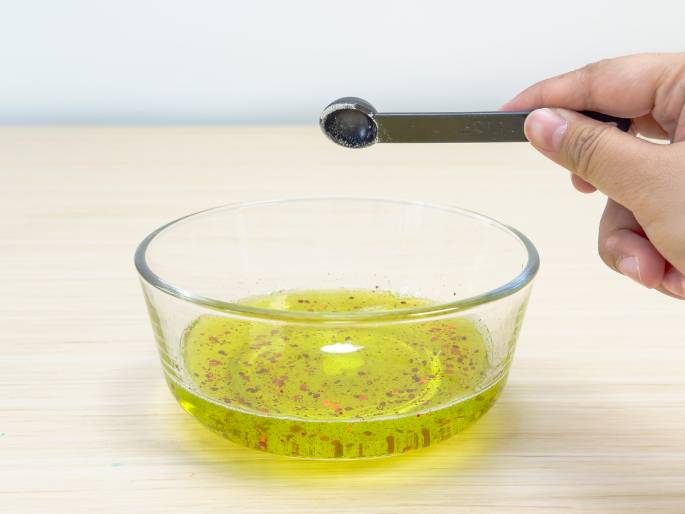 A hand holds a small, black measuring spoon containing a white powder above a clear glass bowl. The bowl, resting on a light wood surface, contains a yellowish liquid mixed with red glitter. The hand appears to be in the process of adding the white powder to the bowl's contents. This image likely depicts a step in a science experiment or the creation of a homemade slime or similar substance.