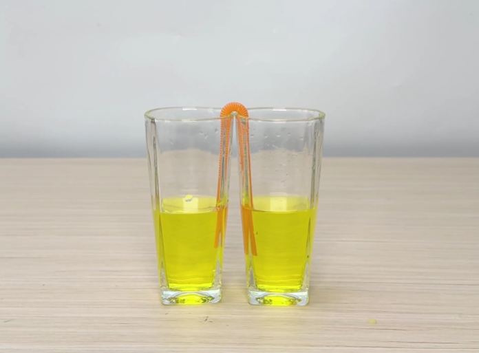 Two clear glasses, partially filled with a bright yellow liquid, sit side-by-side on a wooden table. A flexible orange drinking straw is placed so that one end is submerged in the liquid of each glass. The video appears to be demonstrating a simple science experiment, possibly illustrating the principles of siphoning or capillary action.