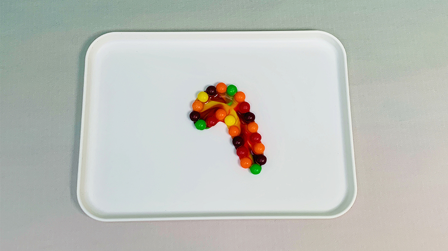 Multicoloured Skittles, arranged in a curved line resembling a candy cane or walking stick, are dissolving on a white tray, their colours bleeding slightly.