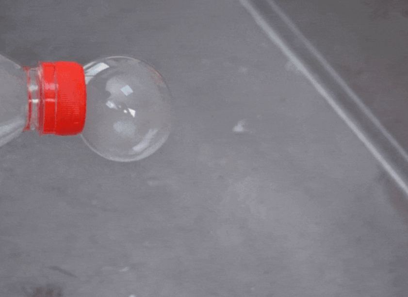 A hand holds a clear plastic bottle with a red cap over a gray surface. The bottle contains a white material, possibly a cloth or paper. A large, clear bubble slowly inflates from the bottle's opening, expanding to nearly the size of the bottle itself. The background is a plain, gray surface, likely a tray or table. This video appears to showcase a science experiment or DIY project involving creating bubbles, possibly using dry ice or a chemical reaction.