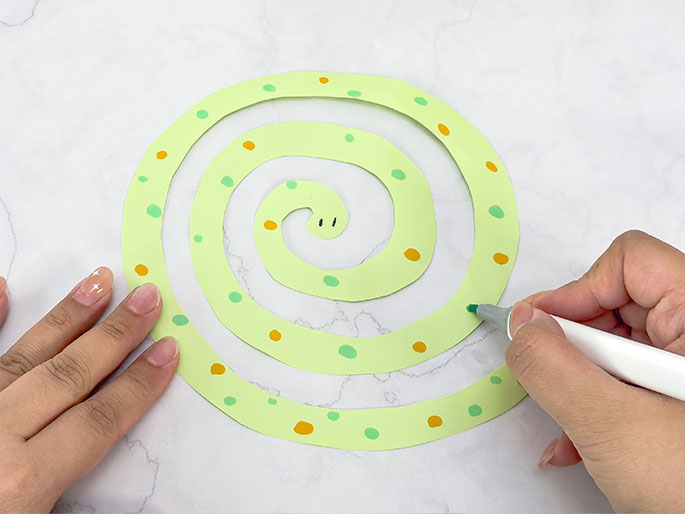 Two hands are shown interacting with a light green spiral cut from paper. One hand holds the spiral flat while the other hand draws small green dots along the spiral's edge with a green marker. Orange dots are already present on the spiral. The spiral has a small, simple face drawn at the center, giving it the appearance of a snake. The background is a marbled surface.