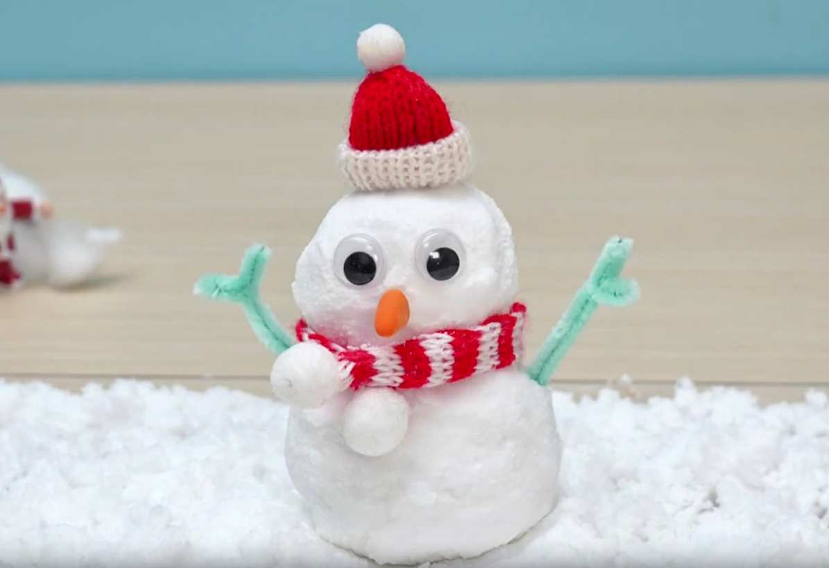 A charming snowman sits on a bed of fluffy white snow against a light blue backdrop. The snowman is adorned with a red and white knitted hat and scarf, googly eyes, an orange carrot nose, light green twig arms, and small white snowball hands. A miniature Santa Claus figure is faintly visible in the background, slightly out of focus. A wooden surface is also visible beneath the snow.