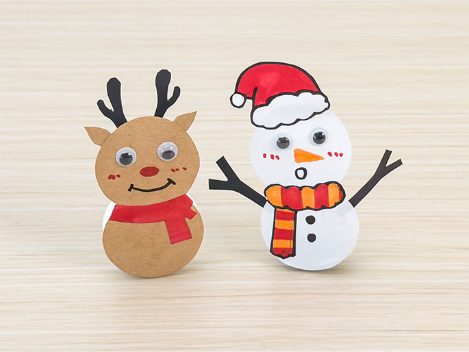 Two charming, finished Christmas crafts stand on a light wood-grain surface. On the left, a reindeer figure is constructed from light brown card stock, featuring googly eyes, a drawn red nose and smiling mouth, black card stock antlers, and a red paper scarf. On the right, a snowman is made of white card stock with drawn features: googly eyes, an orange carrot nose, rosy cheeks, a surprised o shaped mouth, and black buttons. The snowman wears a striped orange and yellow scarf, a red paper Santa hat, and has black card stock arms. These simple crafts evoke a festive holiday spirit.
