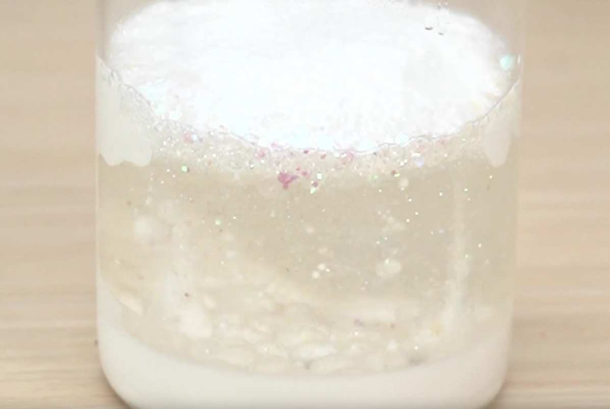 A clear glass jar sits on a light wooden table. The jar contains a layered mixture: a white, opaque substance at the bottom, a clear liquid layer in the middle, and a thicker, white substance at the top. Some of the top layer drips down into the clear liquid, creating a visually interesting effect. Small, sparkling particles are visible within the clear liquid layer. The video showcases the mixing or layering of different substances, possibly a science experiment or the creation of a sensory toy.