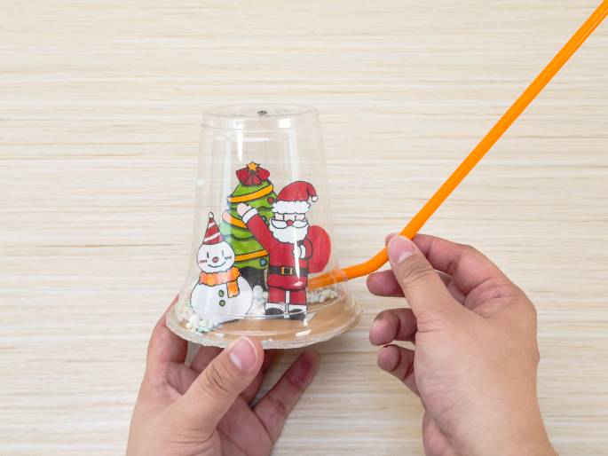 A homemade Christmas snow globe, crafted from a clear plastic cup, sits on a wooden table. Inside the cup, cartoon images of Santa Claus, a snowman, and a decorated Christmas tree are visible. Small, colorful beads, representing snow, are scattered around the figures. An orange straw is inserted into a hole near the bottom of the cup. The video showcases a festive DIY craft project, suitable for children during the holiday season.