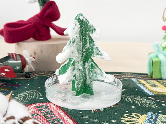 A small, green, cardboard Christmas tree, covered in white, crystalline material, stands in a clear dish filled with the same material, on a green and red Christmas-themed cloth, with other festive decorations, including a red and white toy airplane and a green present, in the background.