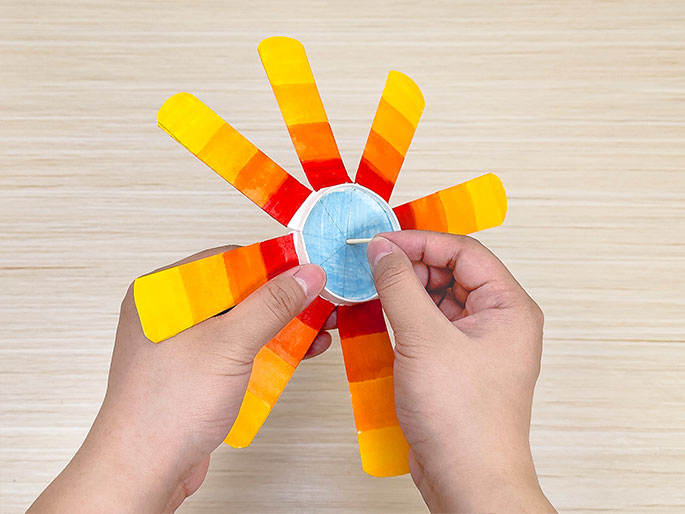 Two hands hold a paper cup flower craft against a light wooden background. The flower, made from a cup cut into segments and colored in shades of yellow, orange, and red, has a light blue center. One hand holds the flower while the other inserts a small wooden stick into the center, likely to create a stem or spinning mechanism.
