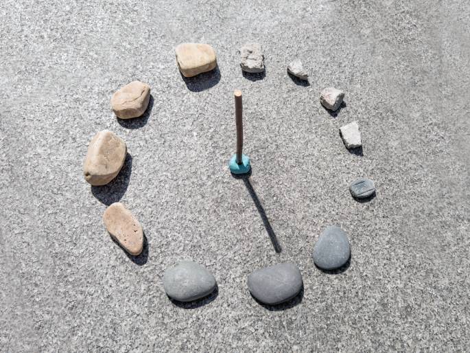 A makeshift sundial is depicted on a textured gray surface, likely concrete or asphalt. A vertical stick, fixed to a light blue base, casts a shadow that falls across the surface. Various rocks and broken pieces of concrete are arranged in a circle around the stick, acting as hour markers. This image demonstrates a simple, DIY sundial.