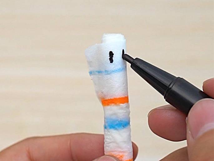 A hand holds a black marker and draws two small vertical black lines onto a white cylindrical object wrapped with orange and blue stripes. The object, possibly made of paper or cloth rolled around a dowel, is held by another hand, partially visible at the bottom of the frame. This image likely depicts a craft project, where details are being added to a handmade item.