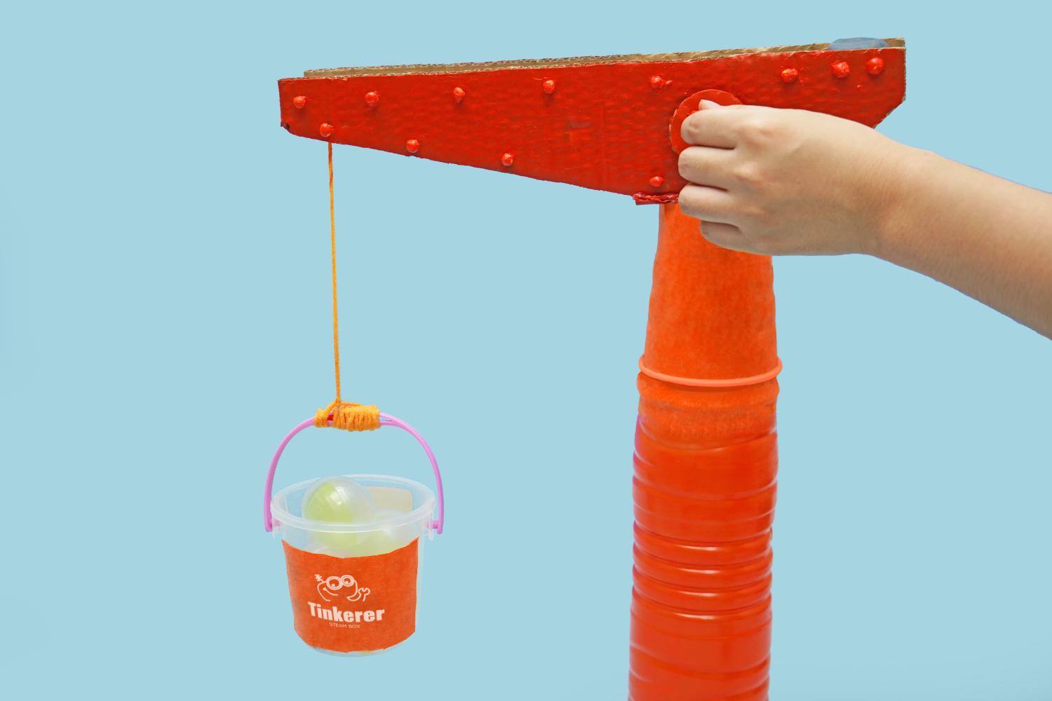 A hand operates a small, painted red cardboard crane, lifting a bucket containing translucent green objects via an orange string, against a pale blue background.