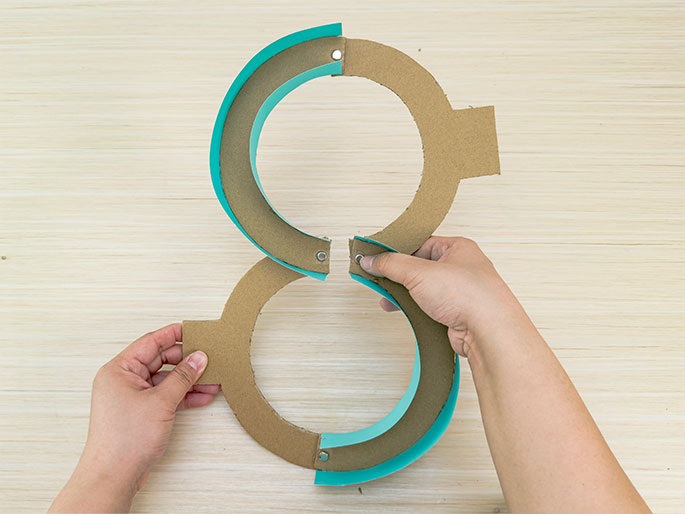 Two hands are assembling a figure-eight shape made of cardboard rings and teal paper strips on a light wood-grained background. Metal brads or fasteners are visible at the points where the teal strips overlap the cardboard, securing them together. The hands are joining the two loops of the eight at the central intersection.