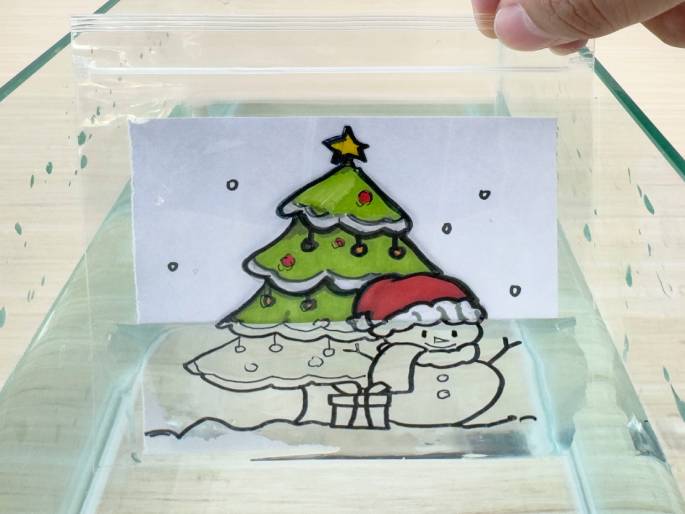 A hand holds a sealed clear plastic bag containing a drawing of a Christmas scene. The drawing features a decorated Christmas tree, a snowman wearing a Santa hat and scarf, and a present, all against a snowy backdrop. The bag is partially submerged in a clear container filled with water. This image likely depicts a step in a craft project, perhaps creating a floating Christmas decoration.