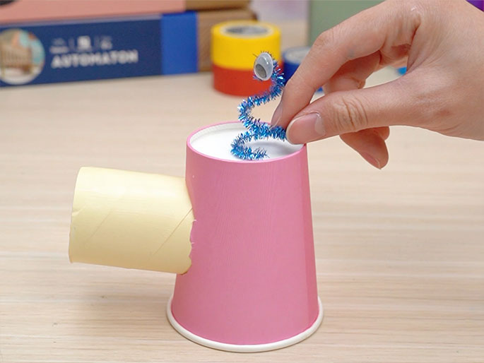 A hand places a sparkly blue pipe cleaner, adorned with a googly eye, into a pink paper cup. The cup has a cardboard tube attached to its side. Rolls of colorful tape and what appears to be a project box labeled AUTOMATON are visible in the background, slightly out of focus. This image depicts a stage in a crafting process, likely for creating a simple automaton or kinetic toy. The light woodgrain surface serves as the backdrop.