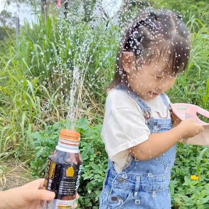 A small child in denim overalls stands in a grassy area getting sprayed with water from a modified can of Wonda coffee held by another person, out of frame.