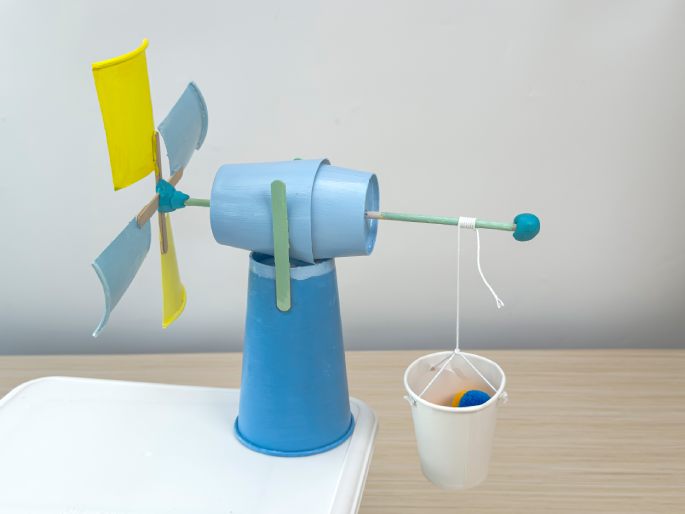 A finished, painted windmill craft project stands on a light-blue paper cup base, which is placed on a larger white square surface. The windmill's tower and horizontal arm mechanism are constructed from light-blue and flattened light-green-painted paper cups, connected by wooden craft sticks. A small ball of teal-colored putty or clay is affixed to the end of the horizontal arm. A string is tied to this teal putty and hangs down to a small, white paper cup bucket with string handles. The bucket contains small yellow and blue balls. The windmill's blades are made from flattened paper cups painted light blue and yellow, attached to crossed craft sticks, and affixed to the horizontal arm with teal putty. This image showcases a completed and colorful DIY windmill model with a loaded bucket.