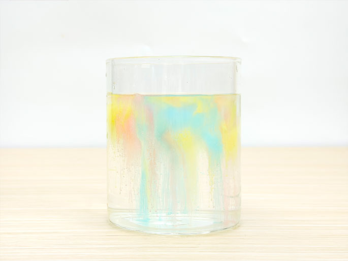 A clear glass sits on a light woodgrain surface, partially filled with water. Swirls of pastel colors—primarily yellow, blue, and pink—are diffused in the water, creating a visually appealing mixture. The colors appear to be slowly mixing and settling, suggesting a recent addition of dye or food coloring. This image likely depicts a science experiment or demonstration involving diffusion or color mixing.