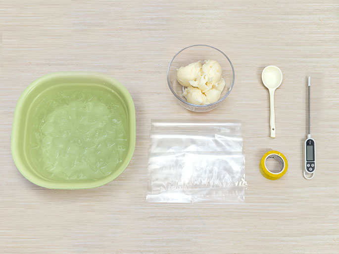 Various items are arranged on a light wooden surface, suggesting preparation for a scientific experiment or cooking activity. A light green bowl filled with ice water is positioned on the left. A small clear bowl containing a white, creamy substance, possibly shortening or butter, is placed above and to the right of the ice water. Next to the creamy substance are a white spoon, a digital thermometer, and a roll of yellow tape. Several clear plastic bags lie flat between the ice water and the other items. The image provides a top-down view of these materials.