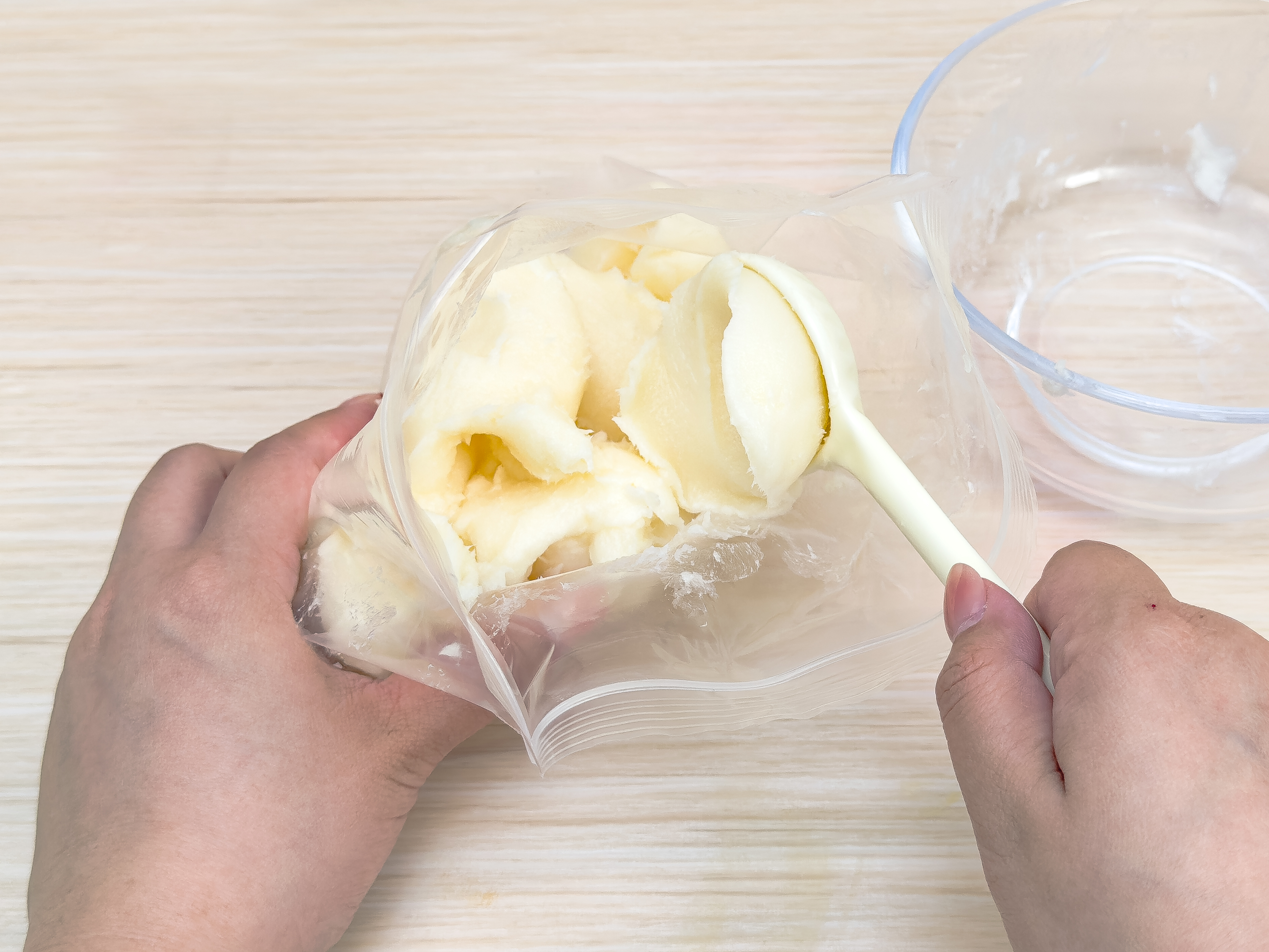 Two hands are shown in the process of transferring a soft, off-white substance, likely shortening or butter, from a clear bowl into a clear plastic zip-top bag. One hand holds the bag open while the other uses a cream-colored spoon to scoop the substance. The background is a light wood surface. The image suggests a food preparation step, possibly for baking or a science experiment.