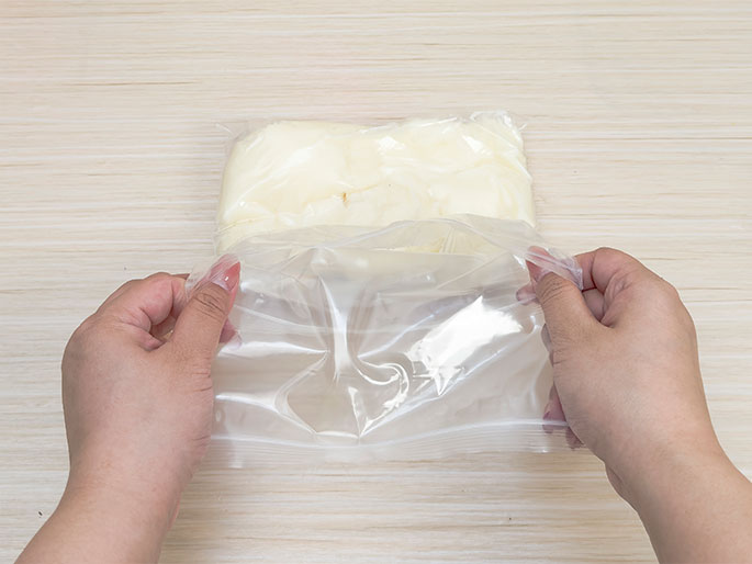 Two hands are flattening a sealed clear plastic zip-top bag containing a creamy, off-white substance, likely shortening or butter. The bag rests on a light wood surface. This image suggests a food preparation step, possibly for baking or a science experiment.