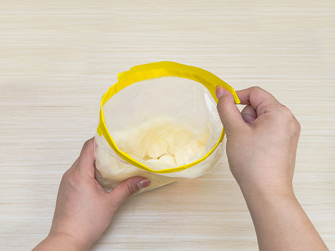 Two hands hold open a clear plastic bag with a bright yellow zip-top seal. Inside the bag is a creamy, off-white substance, likely shortening or butter. The bag is held above a light wood surface. The image suggests a stage in food preparation.