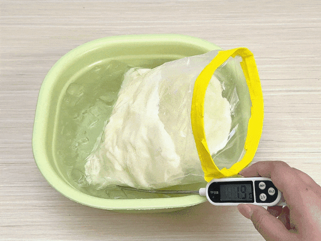 A clear plastic zip-top bag with a yellow seal, containing a soft, off-white substance, is partially submerged in a pale green bowl of ice water. A hand holds a digital thermometer with its probe inserted into the bag. The thermometer displays a temperature of 19 degrees Celsius. The process depicted is likely cooling or chilling a food item. The background is a light wooden surface.