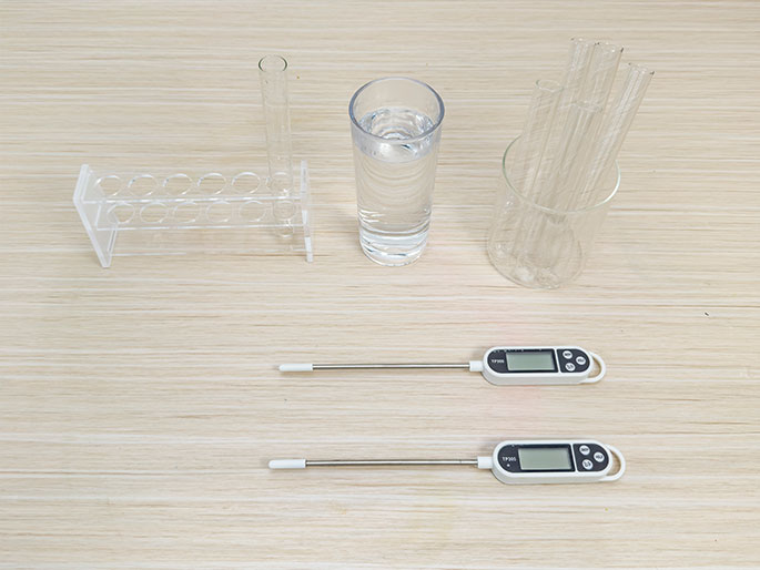 Arranged on a light wood-grain table are various laboratory items. These include a clear acrylic test tube rack with one test tube, a clear glass filled with water, a clear beaker containing several thin glass tubes or rods, and two digital thermometers with long metal probes and white plastic bodies. The items appear to be set up for a scientific experiment or demonstration.