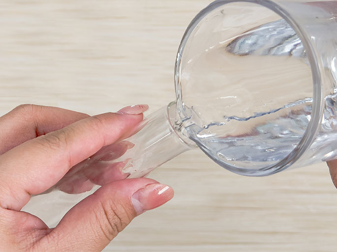 A hand holds a clear glass test tube as water is poured into it from a clear glass. The water is flowing from the glass into the test tube against a light wood-grain background.
