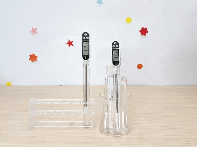 Two digital thermometers are shown against a white background decorated with colorful star and flower stickers. One thermometer stands in a clear plastic test tube rack, and the other is inside a clear glass holding several test tubes. The thermometers display slightly different temperature readings in Celsius. The video subtly zooms in on the thermometers and then back out. The setup suggests a simple science experiment, possibly related to measuring temperature changes.