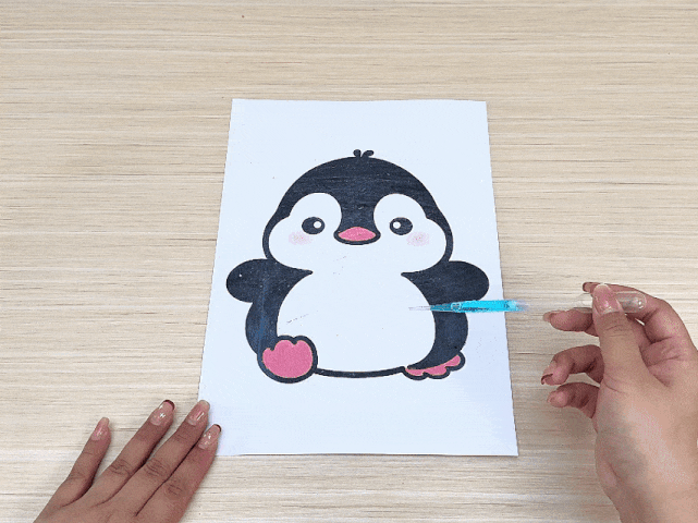 A hand uses a dropper to apply small drops of light blue liquid onto a drawing of a penguin. The penguin is outlined in black on white paper, with pink accents on its cheeks and feet. The blue liquid appears to be spreading slightly on the paper, creating a textured effect. The video focuses on this simple art activity.