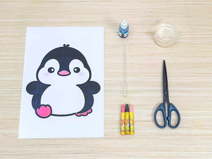 A flat lay on a light wood surface displays craft supplies arranged for a children's activity. The items include a printed image of a cute cartoon penguin on white paper, three crayons (pink, yellow, and dark gray), a pair of blue-handled scissors, a small bottle of blue liquid (likely paint or ink), a clear glass or plastic beaker with a small amount of clear liquid, and a clear plastic pipette or dropper. The arrangement suggests preparation for a penguin-themed craft project.