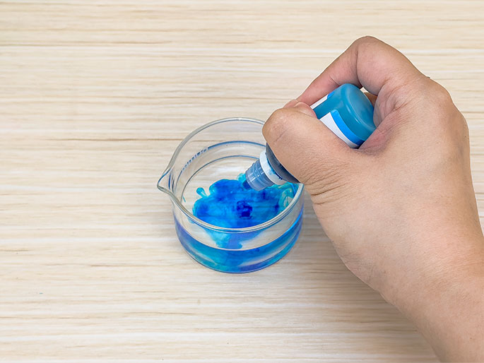 A hand squeezes blue coloring from a small bottle with a nozzle into a clear glass beaker containing a small amount of clear liquid. The blue coloring is swirling and dissolving into the liquid. The beaker sits on a light wood surface.