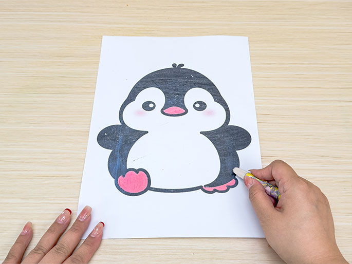A hand holding a gray crayon is coloring a printed image of a cartoon penguin on a piece of white paper. The penguin's body is partially colored gray, and its feet are pink. Another hand is visible at the bottom left corner, holding the paper in place. The background is a light wood surface.
