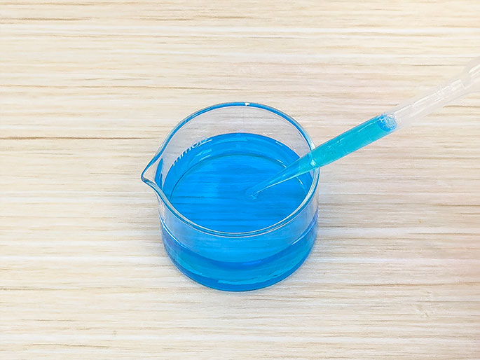 A clear glass beaker filled with bright blue liquid sits on a light wood surface. A clear plastic pipette containing a small amount of the same blue liquid is positioned above the beaker, with the tip just touching the surface of the liquid. This suggests an experiment or activity involving the liquid.
