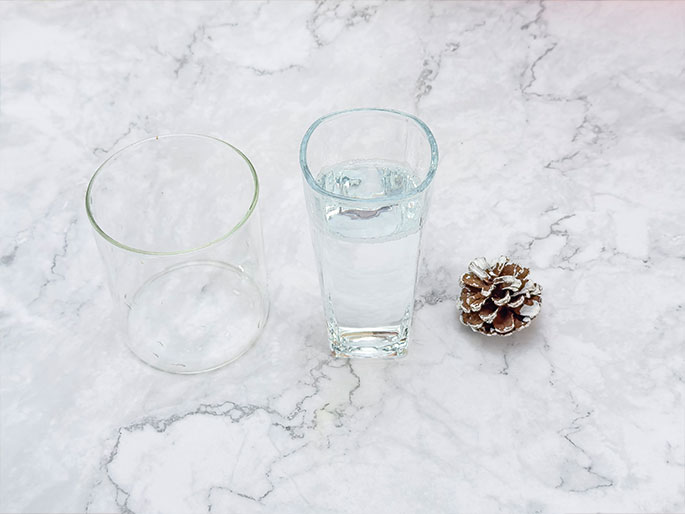 Two clear glasses, one empty and cylindrical, the other partially filled with water and slightly taller with a more rectangular shape, sit on a white marble surface. A small pine cone, dusted with white or silver paint or glitter, lies near the glasses. The arrangement suggests a simple still life or the preparation for a science experiment or demonstration.