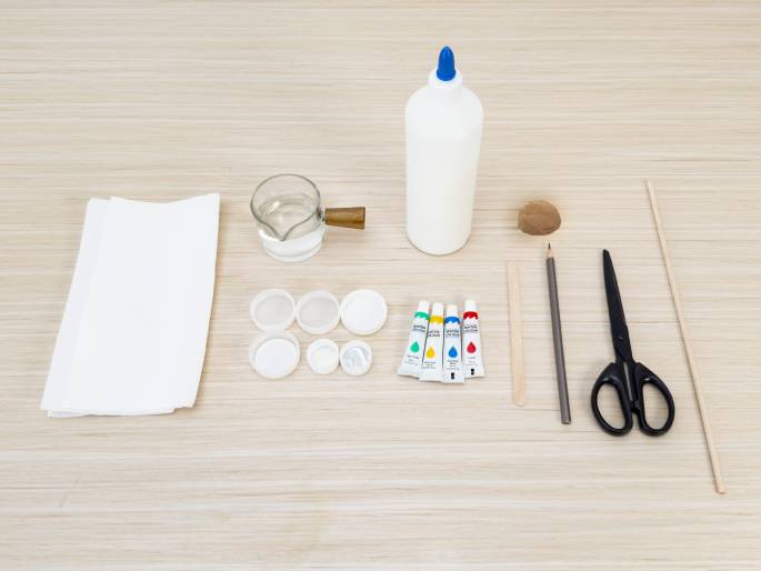 Arranged on a light woodgrain surface are various craft supplies. These include a folded white cloth, a clear glass pitcher with water, a bottle of white glue, a small brown object (possibly a seed pod or nut), four small tubes of paint (green, yellow, red, and blue), a wooden craft stick, a pencil, a pair of scissors, and several small, clear plastic lids. The scene suggests preparation for a children's art or craft activity.