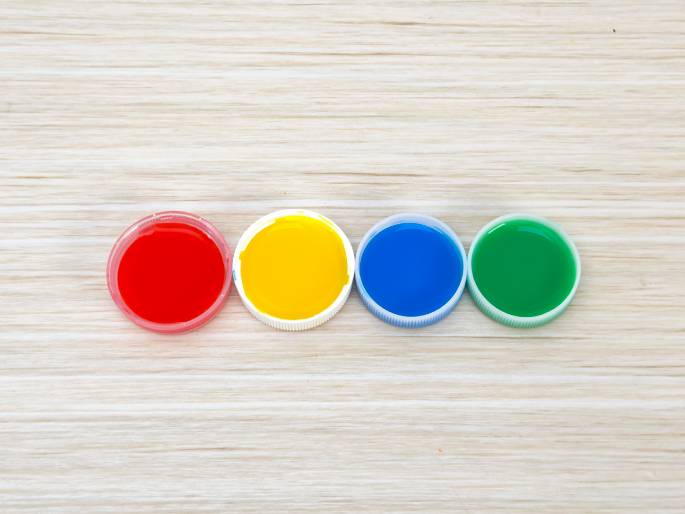 Four plastic bottle caps, each filled with a different vibrant color, are arranged in a row on a light woodgrain table. From left to right, the colors are red, yellow, blue, and green. The image evokes a sense of playful creativity, suggesting an art project or children's activity.