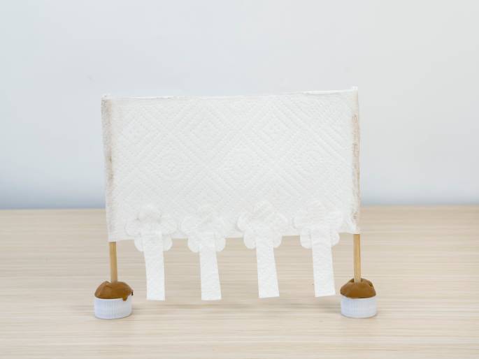 A finished craft project stands on a light woodgrain table against a plain white background. The project consists of a rectangular piece of white textured material, possibly a paper towel, stretched between two thin wooden dowels. Four white flower shapes are glued along the bottom edge, and small white rectangular tabs hang down from each flower. The dowels are inserted into white plastic bottle caps, each filled with brown modeling clay to form a base. The project resembles a small decorative banner or a miniature theatrical backdrop.