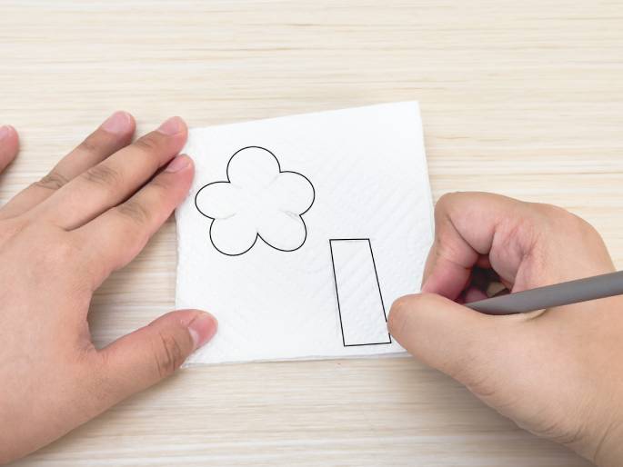 On a light woodgrain table, two hands interact with a white paper napkin. One hand holds the napkin steady while the other hand, holding a pencil, shades within a pre-drawn rectangular shape. Above the rectangle, a simple flower shape is also drawn on the napkin. The scene suggests a crafting activity in progress, possibly creating paper flowers.