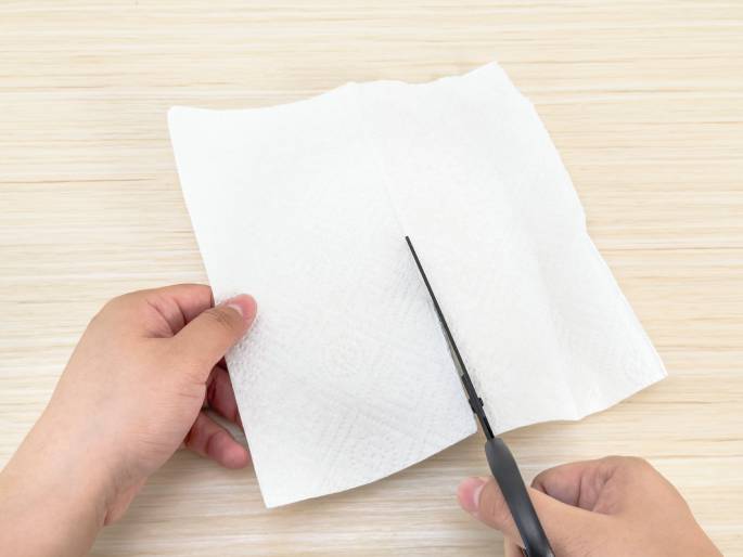 Two hands are positioned on a light woodgrain table, interacting with a white paper napkin. One hand holds the napkin while the other hand uses scissors to cut along the napkin's center crease. The image captures the action of cutting the napkin in half.