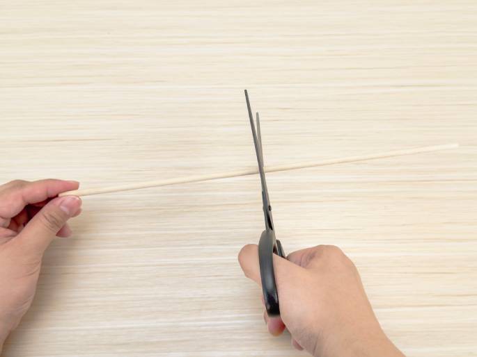 Against a light woodgrain background, two hands are shown. One hand holds a thin, light-colored wooden dowel or stick, while the other hand uses scissors to cut the dowel. The image captures the moment of cutting the wooden stick.