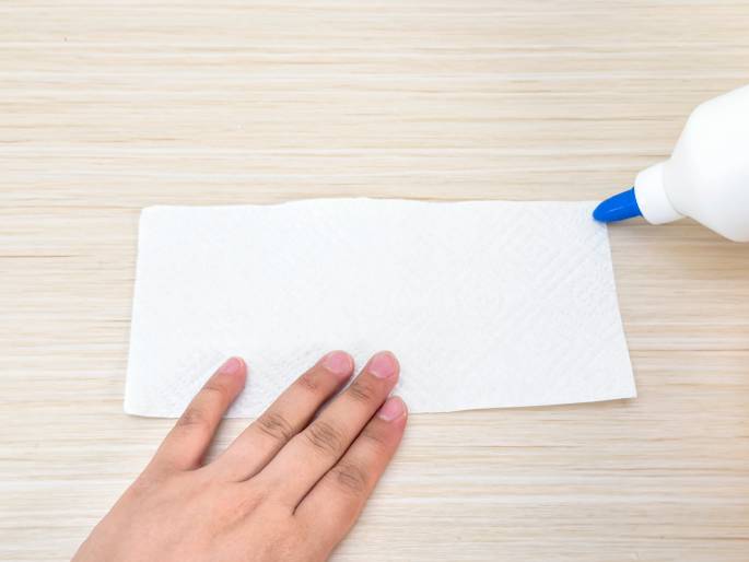 A hand holds a white paper napkin flat on a light woodgrain table. A white glue bottle with a blue tip is positioned at the top right corner of the napkin, seemingly about to apply glue. The image suggests the beginning of a crafting activity involving paper and glue.