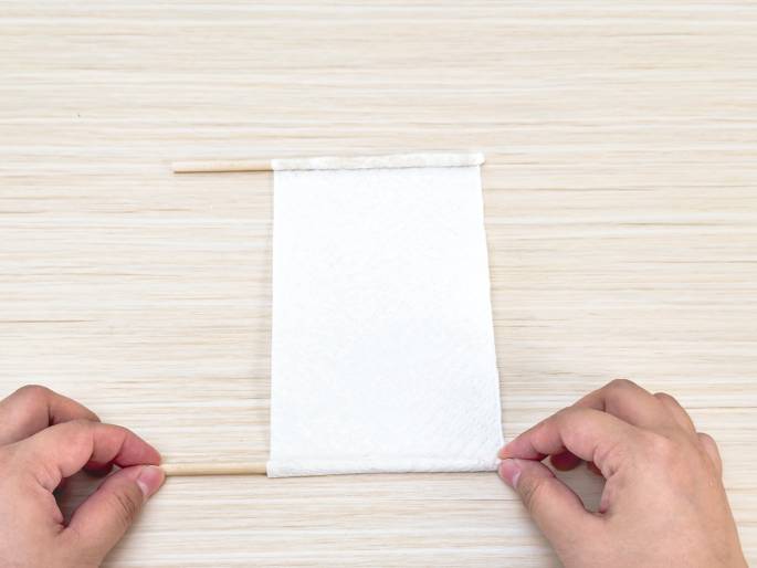 Two hands manipulate a white paper napkin and two thin wooden dowels on a light woodgrain table. The napkin is folded over the dowels, which are positioned at the top and bottom edges of the napkin. The hands appear to be securing the napkin around the dowels, suggesting a crafting process, possibly making a small flag or banner.