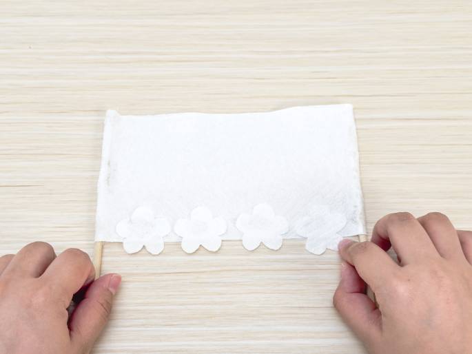 On a light woodgrain table, two hands hold a craft project. A white rectangular piece, likely a napkin or piece of fabric, is stretched between two thin wooden dowels. Glued along the bottom edge of the rectangle are four small, white flower shapes. The hands are holding the edges of the rectangle near the dowels, suggesting the final stages of assembly. The project resembles a small banner or decorative hanging.