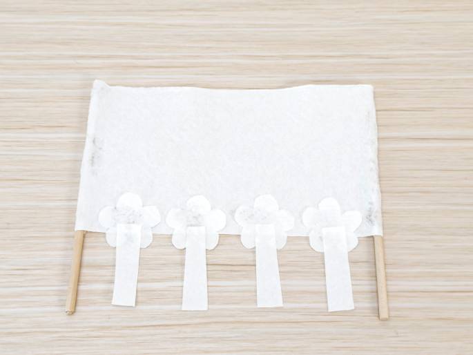 A completed craft project is displayed on a light woodgrain table. A white rectangular piece, possibly a napkin or fabric, is held taut between two thin wooden dowels, forming a small banner. Four white flower shapes are glued across the bottom edge, and hanging down from each flower is a small, white rectangular tab. The overall appearance is delicate and decorative.